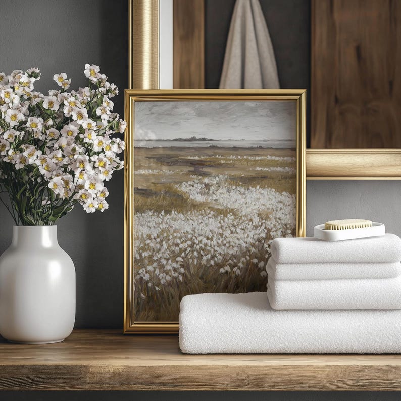 May include: A still-life arrangement featuring a white vase with white flowers, a framed landscape painting, and a stack of white towels. The painting has a gold frame and depicts a field of white flowers. A small white soap dish with a bar of soap is on top of the towels.