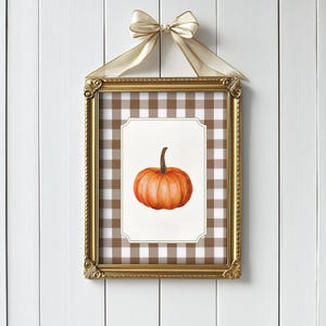 Può includere: Un'opera d'arte incorniciata a tema autunnale con una zucca ad acquerello. La zucca arancione con un gambo marrone è ambientata su uno sfondo bianco all'interno di una cornice dorata ornata. La cornice è sospesa da un nastro color crema.