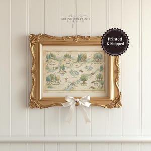 May include: A framed map print with a gold ornate frame, featuring a whimsical illustrated landscape with trees, water, and buildings. A white bow is tied to the bottom of the frame. The text "Printed & Shipped" is on a black circular sticker.