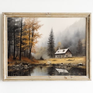 Puede incluir: Una pintura enmarcada de una cabaña en un bosque brumoso. La cabaña es de color marrón claro con un techo blanco y está rodeada de árboles altos. El agua en primer plano refleja los árboles y el cielo.