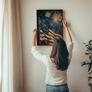 May include: A person hangs a framed painting of an American flag waving in the wind with fireworks in the background. The flag is red, white, and blue. The fireworks are white and yellow.