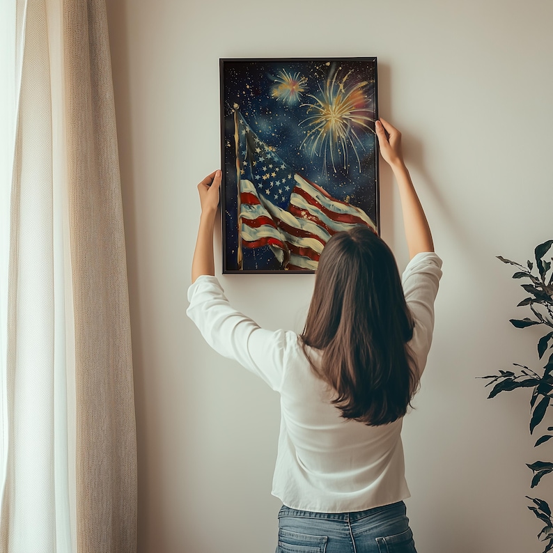 May include: A person hangs a framed painting of an American flag waving in the wind with fireworks in the background. The flag is red, white, and blue. The fireworks are white and yellow.