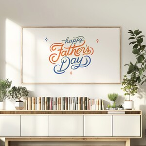 May include: A framed print with the words "happy Father's Day" in a decorative script. The text is in blue and orange, with small star accents. The print hangs above a white cabinet with books and plants.