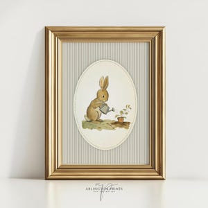 Puede incluir: Una impresión enmarcada con una ilustración en acuarela de un conejo marrón regando una pequeña planta. La obra de arte está enmarcada en un óvalo sobre un fondo a rayas, todo dentro de un marco dorado. La impresión está firmada por Arlington Prints.