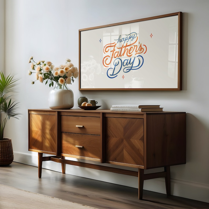 May include: A mid-century modern style wooden sideboard with a framed print that says "Happy Father's Day" in orange and blue script. The sideboard has two doors, two drawers, and a geometric design on the door panels. A vase of flowers and decorative items sit on top.