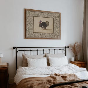 May include: A framed vintage print of a turkey hangs above a bed with a black metal frame. The print has a floral border and a turkey illustration. The bed has white linens, beige pillows, and a brown blanket.