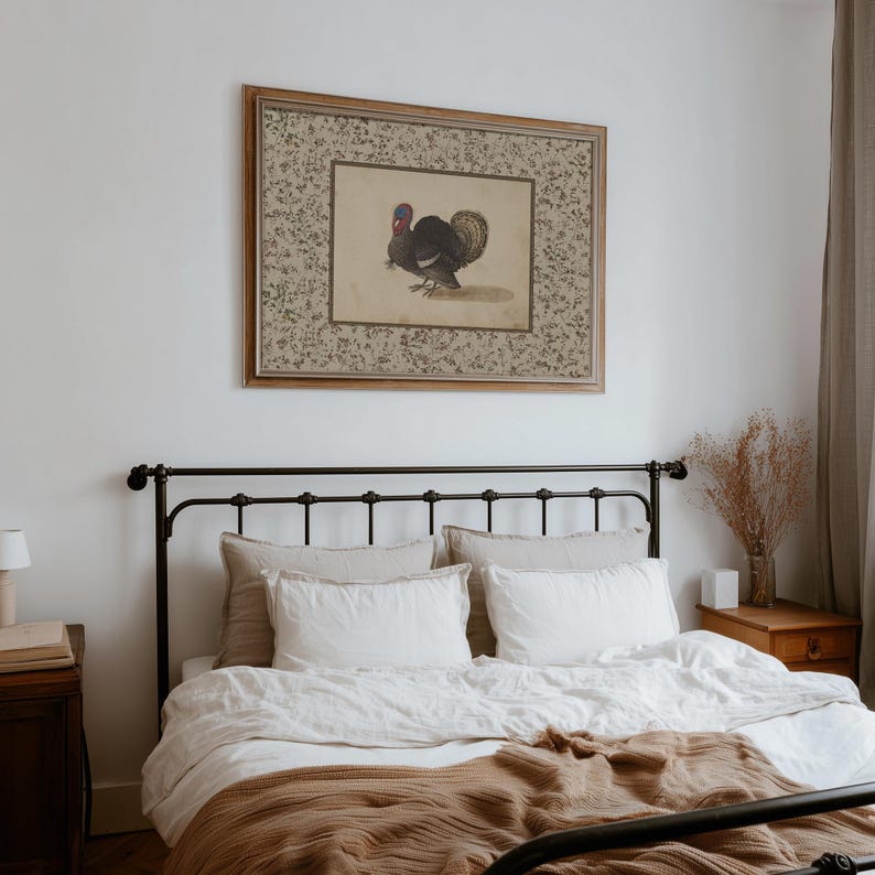 May include: A framed vintage print of a turkey hangs above a bed with a black metal frame. The print has a floral border and a turkey illustration. The bed has white linens, beige pillows, and a brown blanket.