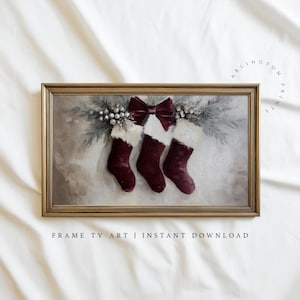 May include: A framed artwork featuring three burgundy Christmas stockings with white fur trim, hanging from a decorative arrangement with a burgundy bow. The artwork is framed in a gold-colored frame. Text at the bottom reads "FRAME TV ART | INSTANT DOWNLOAD".