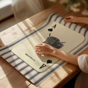 May include: A cream-coloured placemat in the shape of the Ace of Spades playing card. It features a black and white yak's head image and the text "MADE IN THE U.S.A." The placemat rests on a blue and white striped runner.