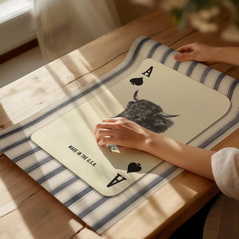 May include: A cream-coloured placemat in the shape of the Ace of Spades playing card. It features a black and white yak's head image and the text "MADE IN THE U.S.A." The placemat rests on a blue and white striped runner.