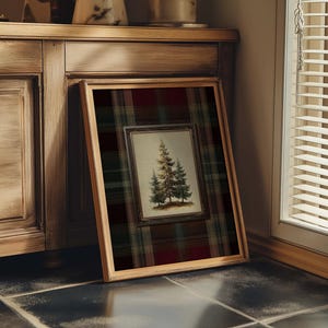 May include: A framed artwork featuring a detailed illustration of evergreen trees. The artwork is set against a plaid background of red, green, and black. The frame is a light brown color, and the artwork is leaning against a wooden cabinet.