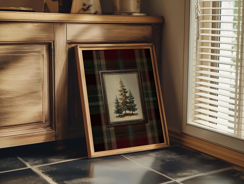 May include: A framed artwork featuring a detailed illustration of evergreen trees. The artwork is set against a plaid background of red, green, and black. The frame is a light brown color, and the artwork is leaning against a wooden cabinet.