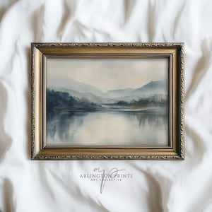 May include: A framed art print depicting a tranquil lake scene with distant mountains. The artwork uses blue and grey tones, set within a gold-coloured ornate frame. The text "ARLINGTON PRINTS ART COLLECTIVE" is displayed at the bottom.