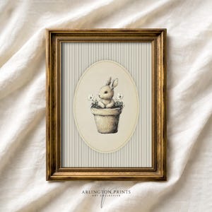 May include: A framed print featuring a charming illustration of a bunny in a flower pot. The artwork is set against a striped background within an oval frame, all encased in a gold-colored wooden frame. The print is signed by Arlington Prints.