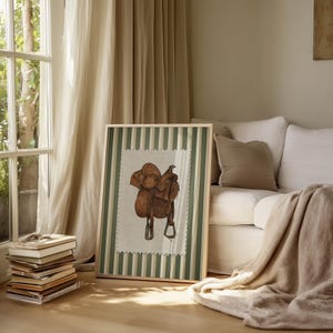 May include: A framed print depicting a watercolour illustration of a brown leather saddle. The artwork has a cream background with a green and white striped border. The frame is on a wooden floor.