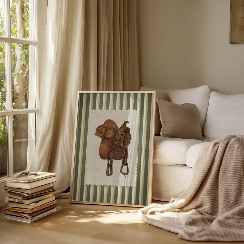 May include: A framed print depicting a watercolour illustration of a brown leather saddle. The artwork has a cream background with a green and white striped border. The frame is on a wooden floor.