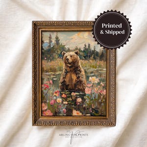 May include: A framed oil painting print featuring a brown bear standing in a pond surrounded by pink and white flowers, with a forest background. The ornate gold frame has a textured, decorative edge. A black circular badge reads Printed & Shipped.