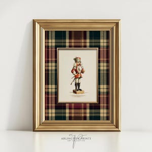 May include: A framed print featuring a vintage illustration of a nutcracker soldier in a red and white uniform, set against a plaid background in shades of green, burgundy, and tan. The frame is gold-colored, and the print is labeled "ARLINGTON PRINTS ART COLLECTIVE."