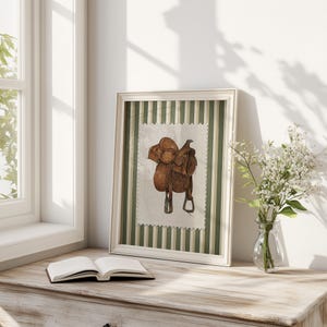 May include: A framed watercolour print of a brown leather saddle. The artwork is set against a green and white striped background and framed in a white wooden frame. The print is displayed on a wooden surface next to an open book and a vase of white flowers.
