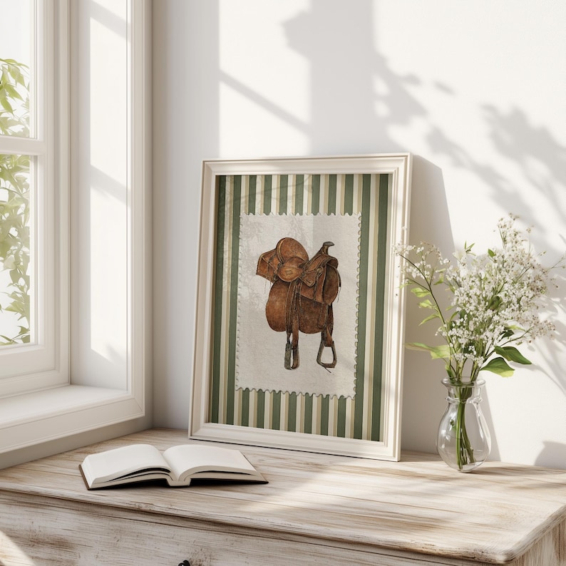 May include: A framed watercolour print of a brown leather saddle. The artwork is set against a green and white striped background and framed in a white wooden frame. The print is displayed on a wooden surface next to an open book and a vase of white flowers.