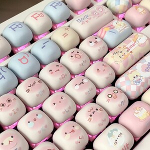 May include: A keyboard with pastel pink, blue, and yellow keycaps featuring cute cartoon faces and Japanese characters. The keycaps are square and have a slightly raised surface.