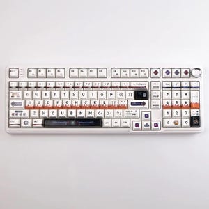 May include: A white mechanical keyboard with custom keycaps. The keyboard features a variety of colors, including orange, purple, and blue. The keys are labeled with letters, numbers, and symbols. The keyboard has a sleek, modern design.
