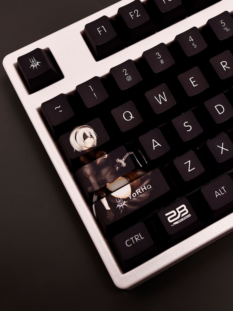 K&ouml;nnte beinhalten: Eine schwarz-wei&szlig;e Tastatur mit benutzerdefinierten Tastenkappen, die einen Charakter aus dem Videospiel "NieR: Automata" zeigen. Die Tastenkappen haben ein schwarz-wei&szlig;es Design mit dem Text "YoRHa" auf einer Taste. Die Tastatur befindet sich auf einer dunkelgrauen Oberfl&auml;che.