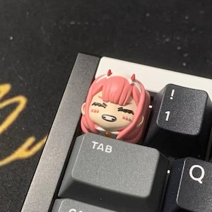 May include: A pink and white anime character keycap with a smiling face and the word "TAB" printed on the keycap.