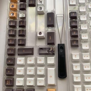 May include: A collection of keyboard keycaps in a clear plastic organizer. The keycaps are a mix of colors, including brown, white, and orange. A black keycap puller is also visible. The keycaps are for a mechanical keyboard.