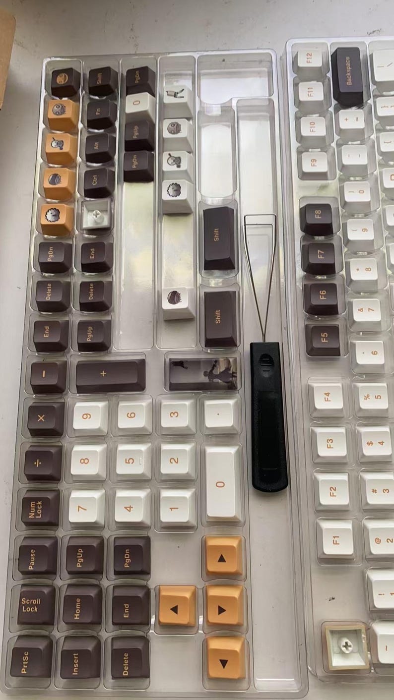 May include: A collection of keyboard keycaps in a clear plastic organizer. The keycaps are a mix of colors, including brown, white, and orange. A black keycap puller is also visible. The keycaps are for a mechanical keyboard.