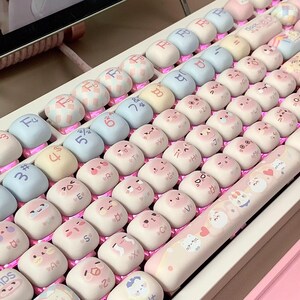 May include: A pink and white keyboard with cute cartoon animal keycaps. The keycaps feature various animals, including cats, dogs, and bunnies. The keyboard also has a pink and white space bar with a heart design.