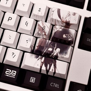 K&ouml;nnte beinhalten: Eine wei&szlig;e mechanische Tastatur mit schwarzen Tastenkappen. Die Tastenkappen zeigen Bilder von Charakteren aus dem Videospiel Nier: Automata. Die Tastenkappen mit Bildern sind mit "Nier" und "Automata" beschriftet.