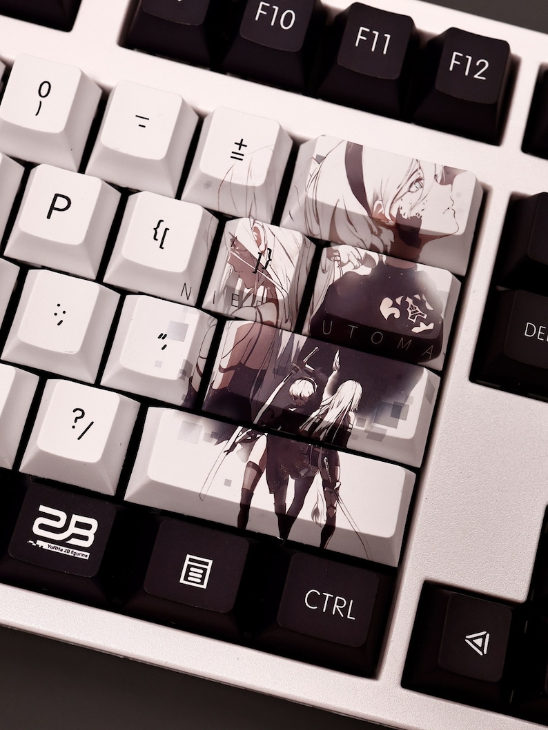 K&ouml;nnte beinhalten: Eine wei&szlig;e mechanische Tastatur mit schwarzen Tastenkappen. Die Tastenkappen zeigen Bilder von Charakteren aus dem Videospiel Nier: Automata. Die Tastenkappen mit Bildern sind mit "Nier" und "Automata" beschriftet.
