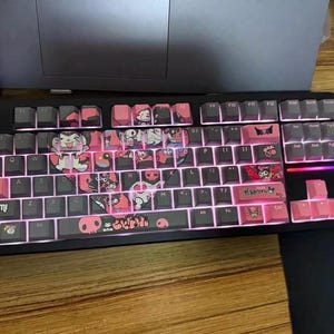 May include: A black mechanical keyboard with pink and white keycaps featuring a cartoon character design. The keyboard has a black frame and a pink and white keycap set with a cartoon character design. The keycaps have a variety of characters, including a cat, a skull, and a heart. The keyboard also has a pink and white space bar with the word "KUROMI" printed on it.