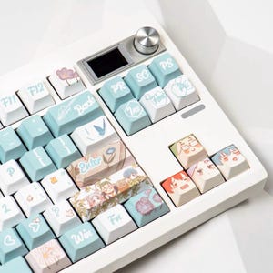Könnte beinhalten: Eine weiße mechanische Tastatur mit individuellen Tastenkappen. Die Tastenkappen sind hellblau und weiß, mit verschiedenen Illustrationen und Text. Die Tastatur hat einen kleinen Bildschirm und einen silbernen Drehregler. Die Tastenkappen zeigen florale und Anime-Designs.