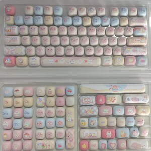 May include: A pastel pink and blue keyboard with cute cartoon characters on each key. The keyboard has a white and pink space bar with a cartoon character on it. The keyboard is on a white surface.