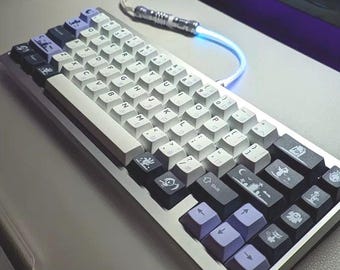 Op de afbeelding: Een wit en grijs mechanisch toetsenbord met aangepaste toetsen met verschillende symbolen en ontwerpen. Het toetsenbord heeft een zilveren frame en een wit en grijs snoer.