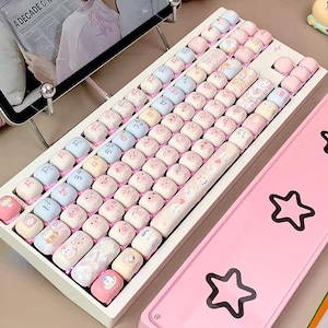 May include: A white keyboard with pink and blue keycaps featuring cartoon animal designs. The keyboard has a white frame and a pink and white cable. The keycaps have a variety of designs, including a cat, a dog, a rabbit, and a bear.