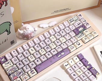 May include: A mechanical keyboard with a light beige frame and custom keycaps featuring purple and white color schemes. The keycaps display various cartoon illustrations, including cats, stars, and moons. A separate numeric keypad is included.