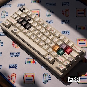 May include: A beige mechanical keyboard with custom keycaps featuring Japanese characters and colorful gaming-themed icons. The keyboard sits on a gaming mousepad with a repeating pattern of game controllers and text. The keycaps include the text "GAME OVER".