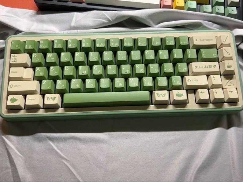 May include: A green and cream keyboard with Japanese characters and symbols. The keyboard has a cream-colored frame and green keys. The keys have white lettering and some have small green symbols. The keyboard has a row of function keys at the top and a row of arrow keys on the right side. The keyboard has a cream-colored space bar with the word "Super" printed on it.