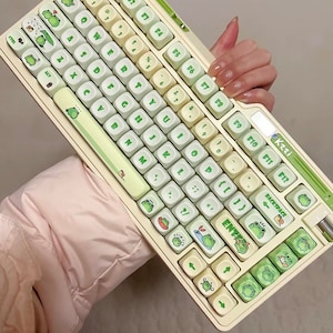 May include: A white and green keyboard with frog-themed keycaps. The keyboard has a number pad and function keys. The keys are labeled with letters, numbers, and symbols. The keyboard has a green frog on the top left corner.