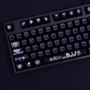 Pode incluir: Um teclado mecânico preto com teclas personalizadas que apresentam personagens e designs de anime. As teclas são brancas com contornos pretos e incluem o texto "Gundam Seed" e "Mobile Suit Gundam UC".