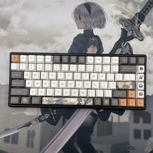 May include: A black mechanical keyboard with white, gray, and orange keycaps. The keyboard features custom keycaps with anime-style artwork and the text "NieR Automata". The background shows a matching anime-style illustration.