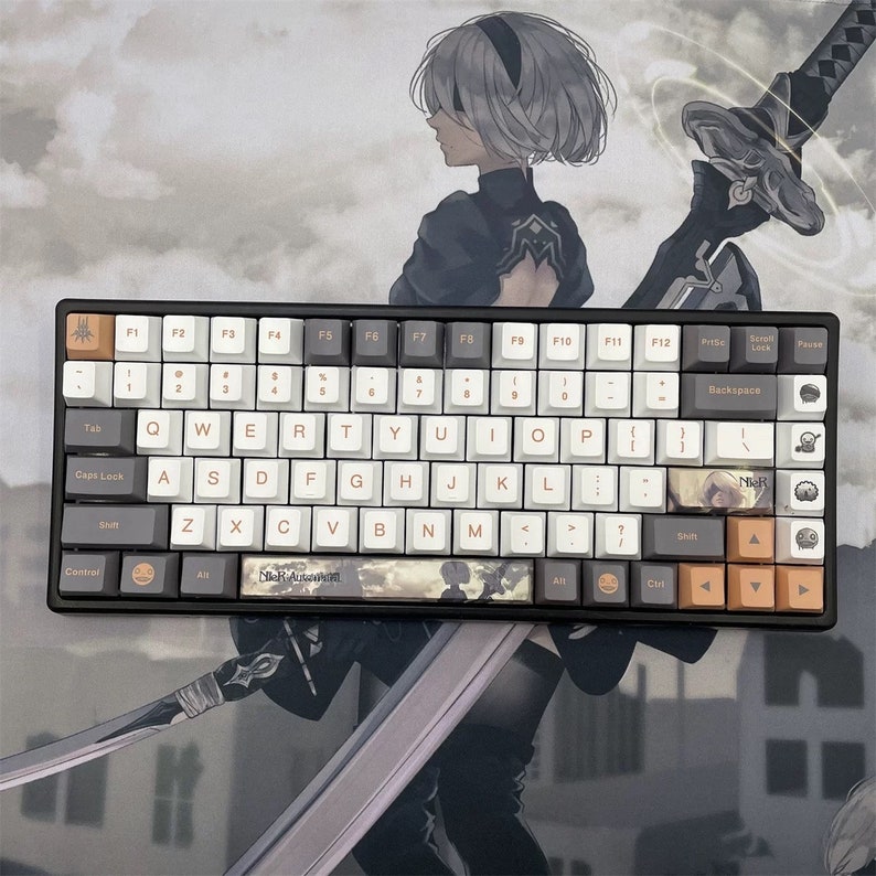 May include: A black mechanical keyboard with white, gray, and orange keycaps. The keyboard features custom keycaps with anime-style artwork and the text "NieR Automata". The background shows a matching anime-style illustration.