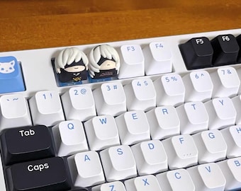 May include: A white mechanical keyboard with custom keycaps. Two keycaps feature anime-style character designs. Other keys are white with blue lettering. A blue keycap with a fox face is also visible. The keyboard sits on a wooden surface.
