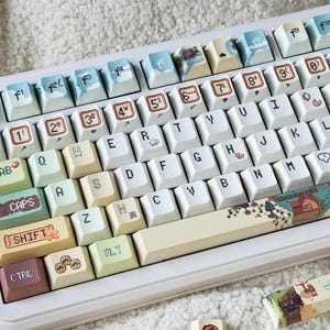 May include: A white mechanical keyboard with pastel-colored keycaps featuring various designs, including a sheep, a house, and a heart. The keyboard has a cream-colored space bar and a light brown bottom row.