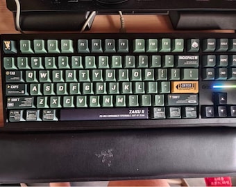 May include: A black mechanical keyboard with green keycaps and a black wrist rest. The keyboard has a label that says "ZAKU II" and "MS-06S COMMANDER TYPE, SORT OF ZION".