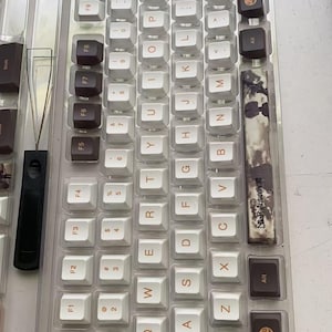 May include: A clear plastic tray holding a set of custom mechanical keyboard keycaps. The keycaps are a mix of white, brown, and black, with some featuring unique designs. A keycap puller is also visible.