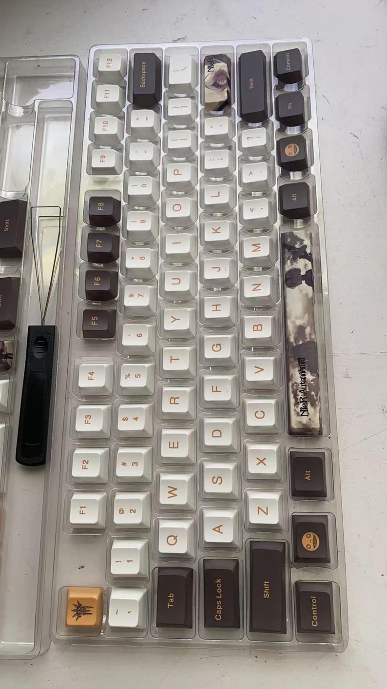 May include: A clear plastic tray holding a set of custom mechanical keyboard keycaps. The keycaps are a mix of white, brown, and black, with some featuring unique designs. A keycap puller is also visible.